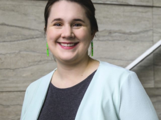 a young woman in a light green blazer and grey dress smiles at the camera while leaning against a railing on a marble staircase