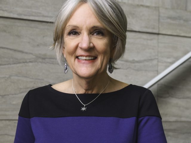 a middle-aged woman in a navy blue and black dress smiles at the camera while leaning against a railing on a marble staircase