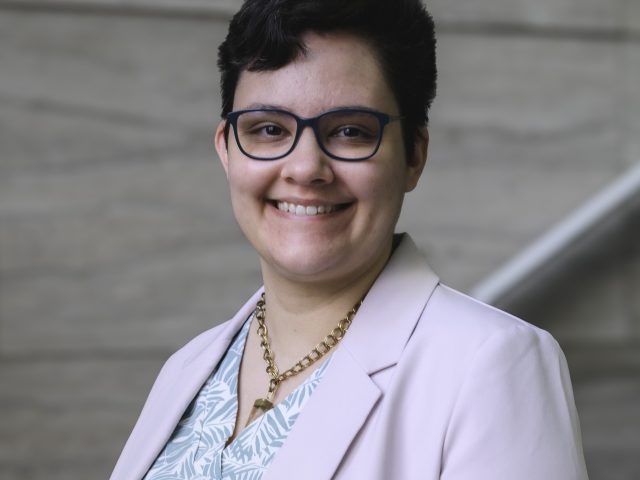 a young woman in a light pink blazer and mint leaf patterned shirt smiles at the camera while leaning against a railing on a marble staircase