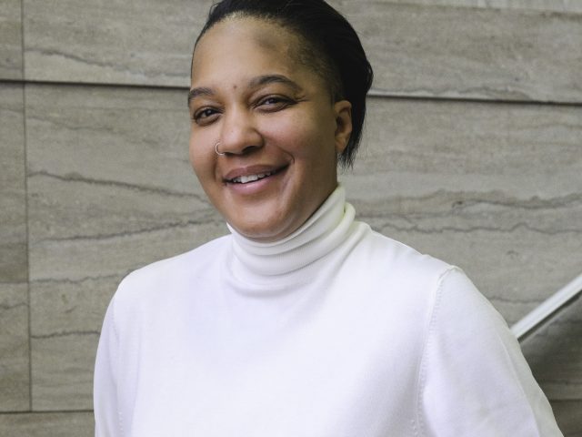 a young woman in a white turtleneck, smiles at the camera while standing in a marble staircase