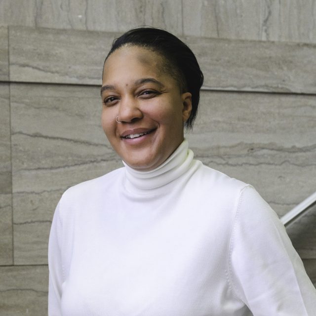 a young woman in a white turtleneck, smiles at the camera while standing in a marble staircase