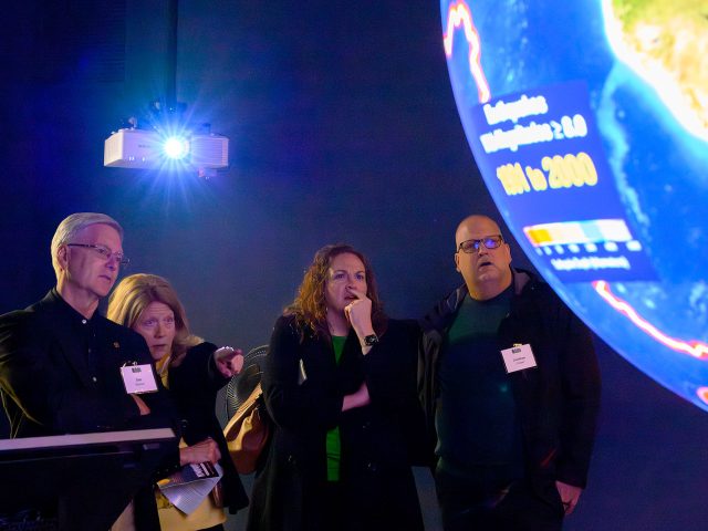 4 people in a dark room looking at a large globe with projected data on it.