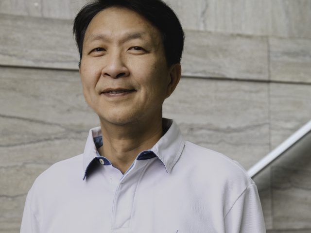 a middle-aged man, in a white collared shirt, smiles at the camera while standing in a marble staircase