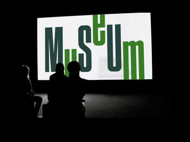 Visitors in a dark room watching an exhibition slideshow with the MSU Museum logo showing on a white screen.