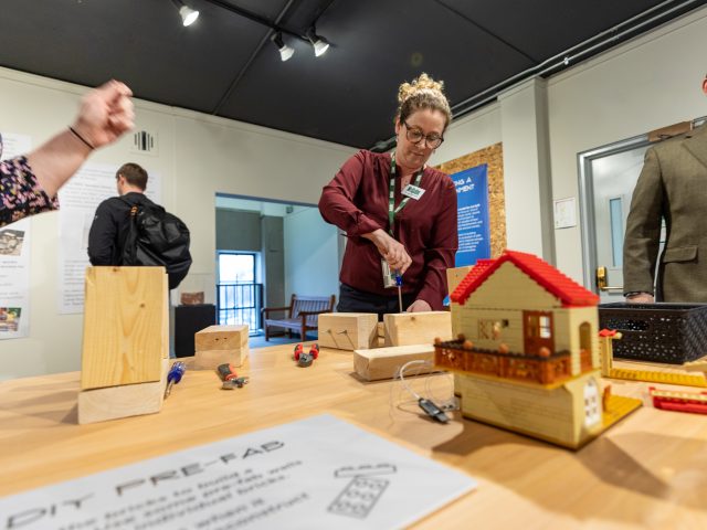 A woman in a burgandy sweater and MSU Museum lanyard demonstrates part of the Domicology exhibit (Teresa Goforth, Director of Exhibitions), with wooden building blocks sitting out on a table as passerby move around her.