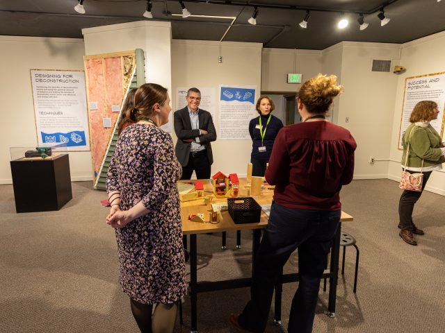 A group stands in the Domicology exhibit, discussing a centerpiece on a table with wood pieces and mini houses.