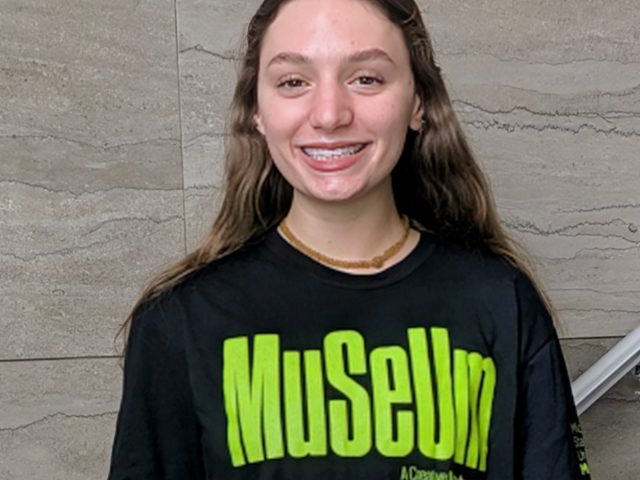 A smiling young woman with long brown hair and braces stands against a gray tiled wall, wearing a black T-shirt with bright green text that reads “Muueum.”
