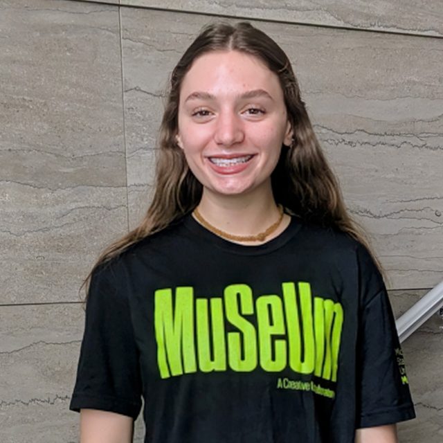 A smiling young woman with long brown hair and braces stands against a gray tiled wall, wearing a black T-shirt with bright green text that reads “Muueum.”
