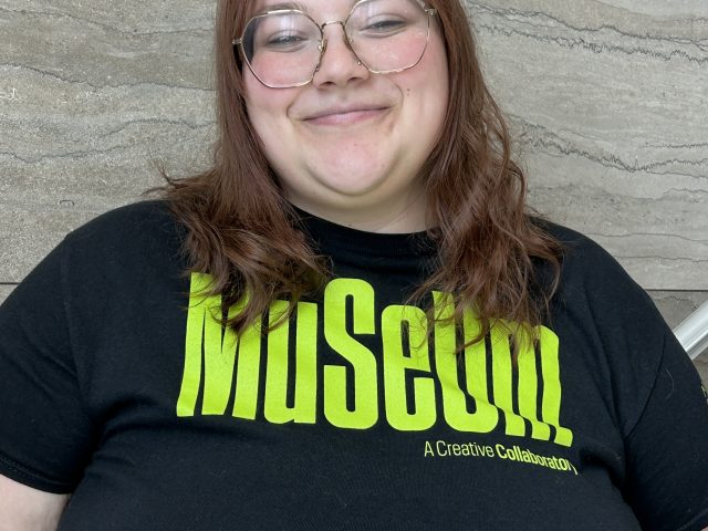 A smiling person with shoulder-length reddish-brown hair and round glasses stands against a textured stone wall, wearing a black T-shirt with bright green “Museum” text and smaller “A Creative Collaboration” lettering.