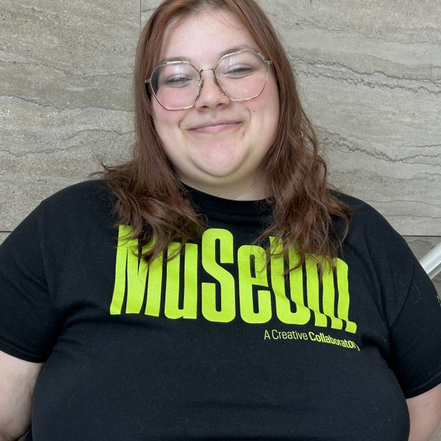 A smiling person with shoulder-length reddish-brown hair and round glasses stands against a textured stone wall, wearing a black T-shirt with bright green “Museum” text and smaller “A Creative Collaboration” lettering.
