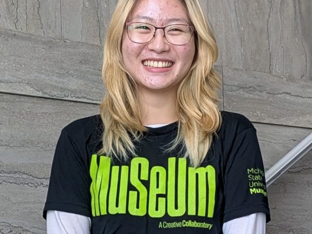 A smiling person with shoulder-length blonde hair and glasses stands against a gray stone wall, wearing a black “Museum” T-shirt over a light gray long-sleeve shirt.
