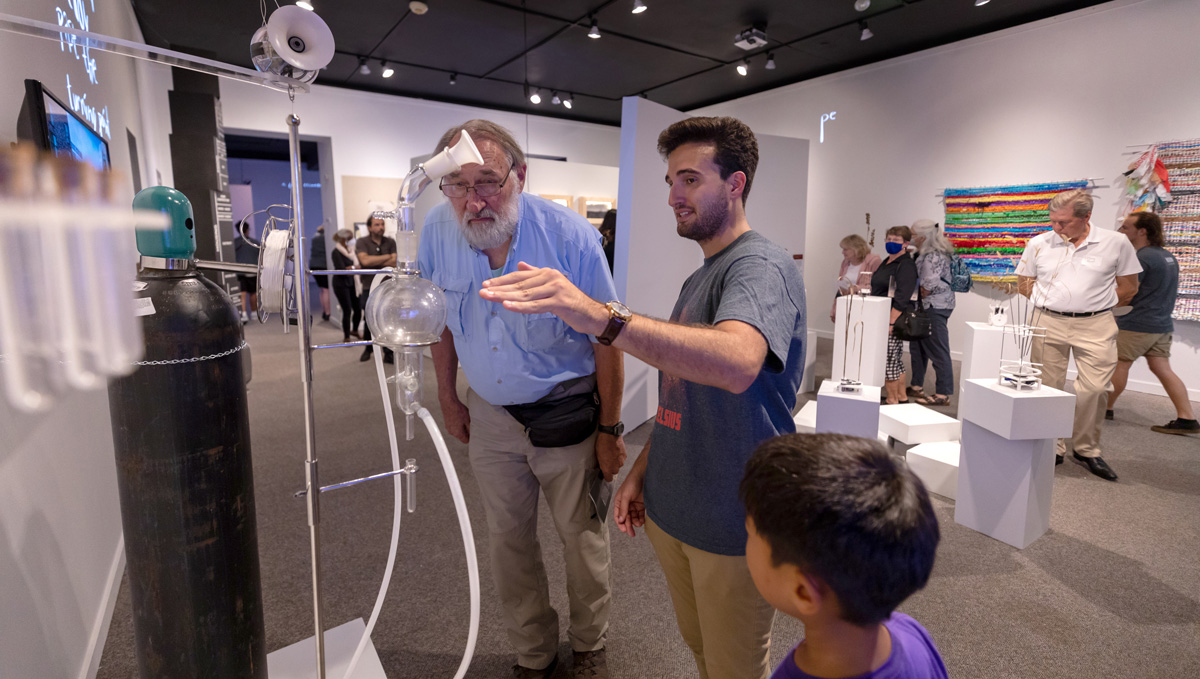 image of and older man, a younger man, and a young boy looking at a museum exhibit. The exhibit has tubes and a large oxygen tank.