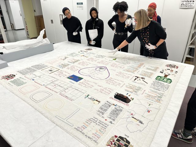 MSU Museum curator and four visitors look at a quilt in the cultural collections area. Quilt features text and images detailing the Todd family history