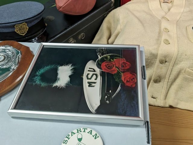 MSU Spartan Marching Band memorabilia on a table, including marching band hat, plaque, picture frame, button, instrument case, football, and cream button-up sweater.
