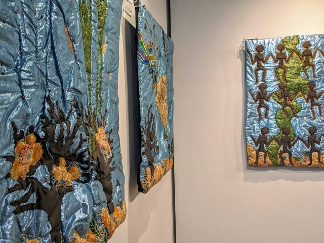 Three quilts made with a shining 3D fabric hang on white walls in an exhibit, depicting topics of Afrofuturism.