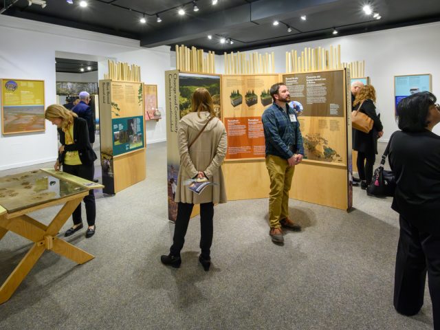 People walk around an exhibit, Knowing Nature, examining wood stands with posters and photos.