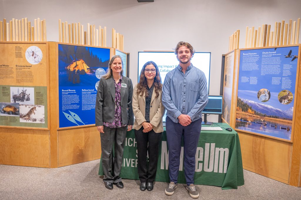 Denice Blair, Director of Education wearing a gray suit, stands next to two MSU students who worked on the RFID project, all three in front of "Knowing Nature" exhibit panels and a table with green MSU Museum tablecloth.
