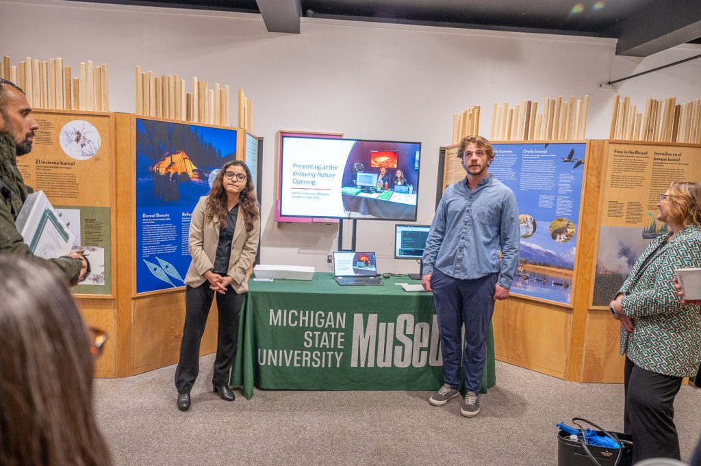 Two MSU students stand at a table with the MSU Museum logo, explaining their work to a crowd.