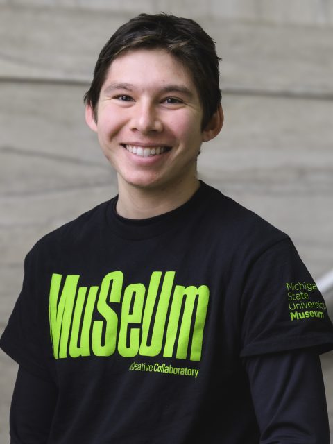 Block image a young man in a black shirt with green lettering that says Museum smiles at the camera while standing in a marble staircase