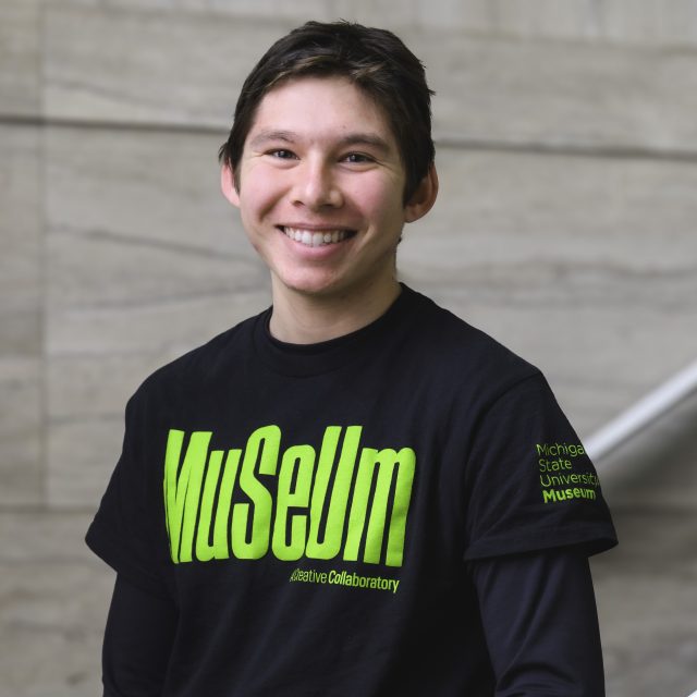 a young man in a black shirt with green lettering that says Museum smiles at the camera while standing in a marble staircase