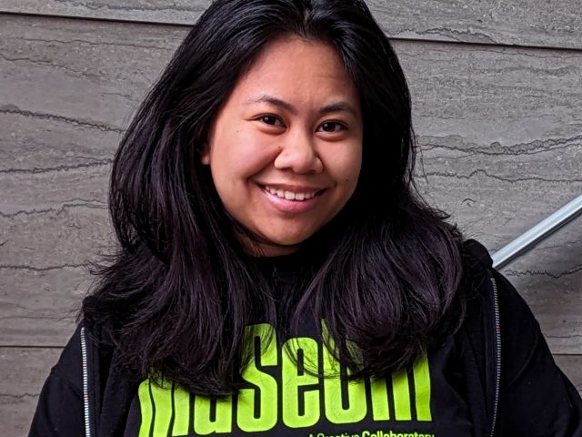 A young woman with shoulder-length black hair wearing a black hoodie and black t-shirt with green text reading "MUSEUM" standing in front of a marble staircase.