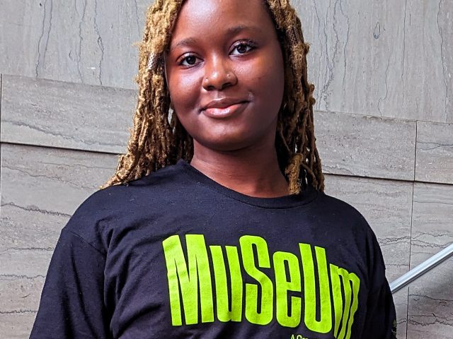 a young black woman with braided light hair wearing a black t-shirt with green text reading "MUSEUM" stands in front of a marble staircase.