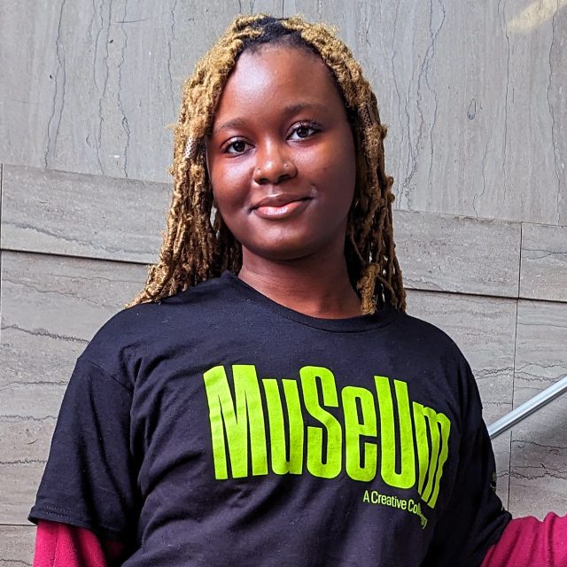 a young black woman with braided light hair wearing a black t-shirt with green text reading "MUSEUM" stands in front of a marble staircase.