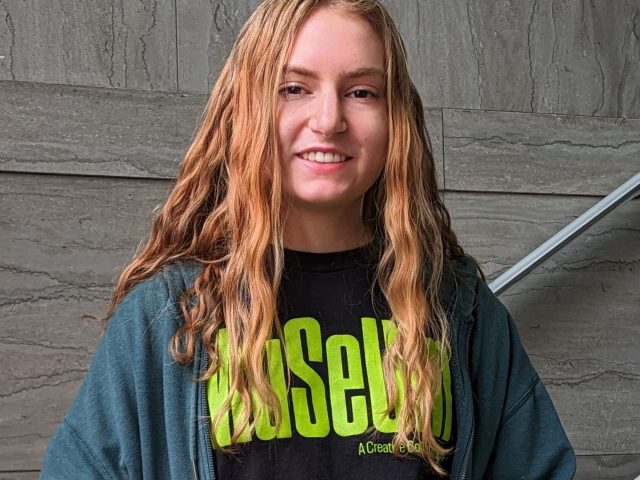 A young woman with curly blonde hair wearing a dark green hoodie and black t-shirt reading "Museum" in green letters stands in front of a marble staircase