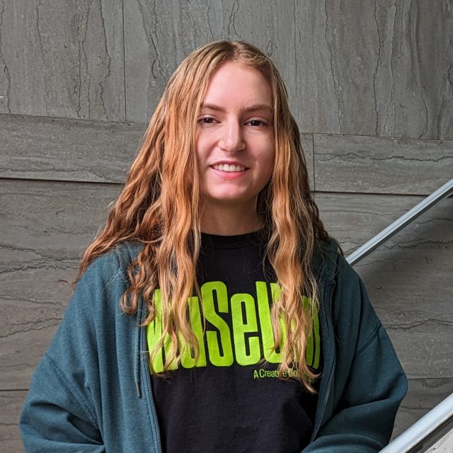 A young woman with curly blonde hair wearing a dark green hoodie and black t-shirt reading "Museum" in green letters stands in front of a marble staircase