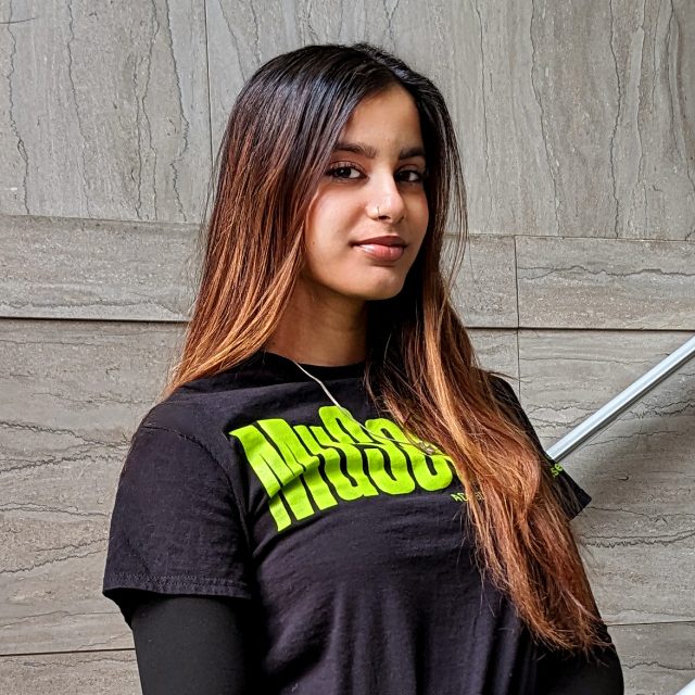 a young woman with long dark hair in a brown ombre wearing a black shirt with green text reading "MUSEUM" standing in front of a marble staircase.
