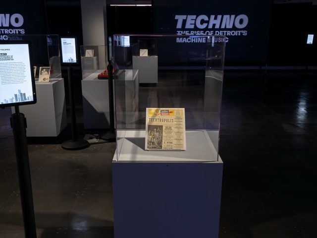 An exhibit display featuring a framed newspaper advertisement for enclosed in a clear glass case on a white pedestal. To the left, an informational digital display provides context about the object. In the background, additional exhibits are visible, along with a large screen displaying the words 