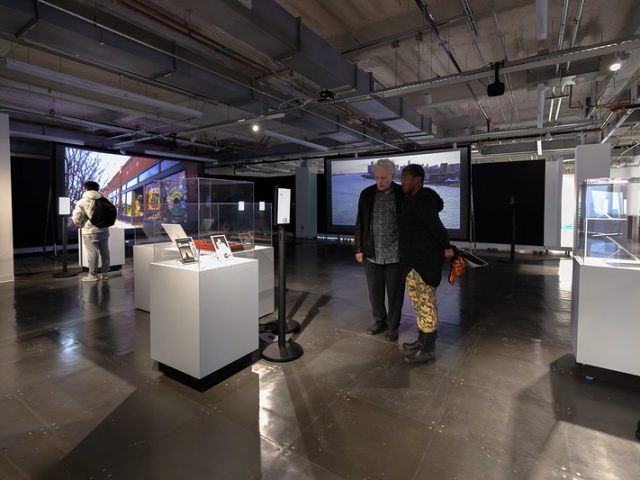 Visitors explore an exhibition space featuring Techno: The Rise of Detroit’s Machine Music. In the foreground, two people stand closely together, engaged in conversation while looking at a display case. To the left, another visitor wearing a white jacket and backpack interacts with a digital information kiosk. Several white pedestals hold artifacts, including books and magazines related to techno music. Large projection screens in the background display images of an urban waterfront and a mural-covered building. The exhibition space has an industrial aesthetic with exposed ceilings and dark flooring, illuminated by spotlights highlighting the displays.