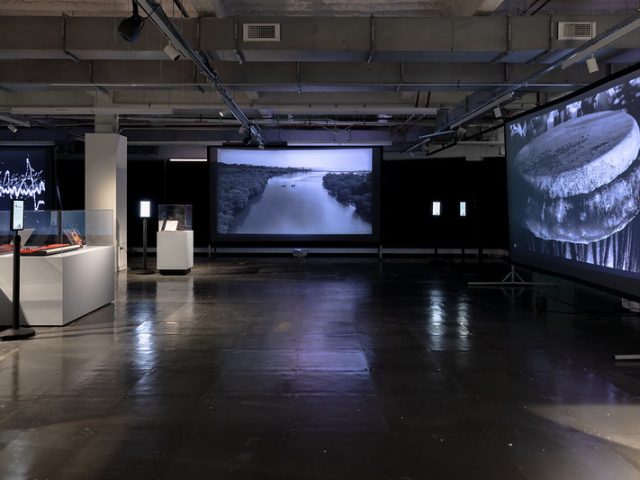 A dimly lit exhibition space featuring large projection screens displaying black-and-white visuals of a river landscape and a close-up of a drum. Various display cases with artifacts, including books and electronic equipment, are positioned throughout the room, accompanied by digital information kiosks. One screen in the background shows an abstract sound wave pattern. The industrial-style setting has exposed ceilings and dark flooring, with spotlights highlighting key exhibits.