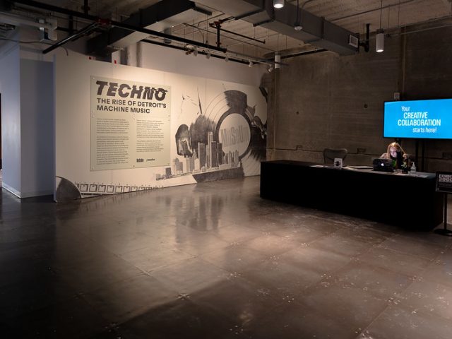 The entrance area of the Techno: The Rise of Detroit’s Machine Music exhibition. A large wall display features the exhibition title along with an introduction text, accompanied by an illustration of a vinyl record, a DJ’s hands, and a city skyline. To the right, a reception desk with a staff member working on a laptop is positioned against a textured concrete wall. A digital screen above the desk displays the message 