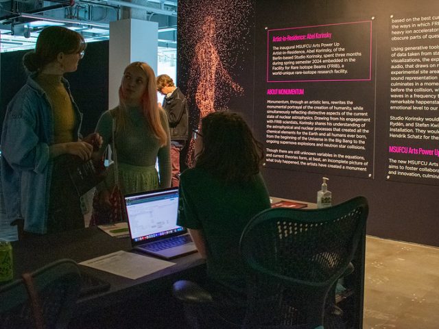 A small group of people gather around a booth at an art exhibit. Two attendees converse with the person seated at a desk, who is using a laptop. Behind them, a large informational panel describes an art installation by Abel Korinsky as part of the MSUFCU Arts Power Up program, with abstract digital artwork displayed nearby.