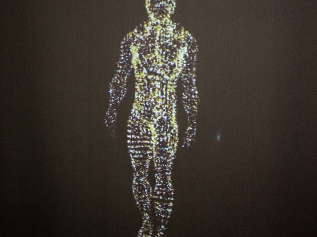 Digital figure of a human body composed of glowing particles, projected against a dark background. The figure appears to be standing or walking, with a futuristic, ethereal quality, as the particles shimmer and form the outline of the body.