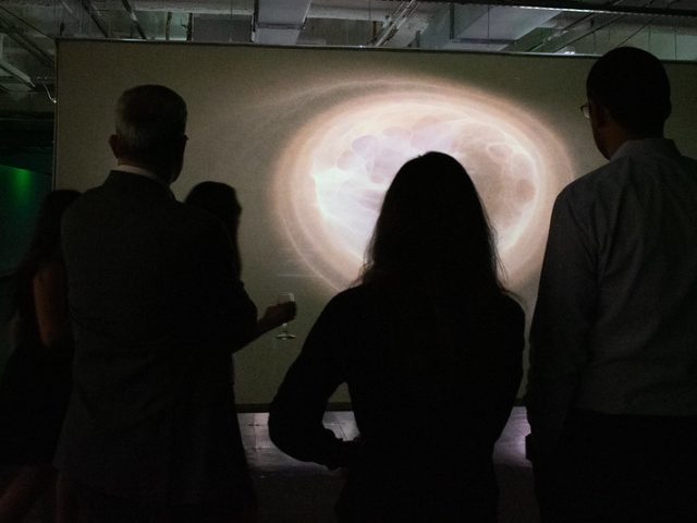 Silhouettes of several people stand closely together, observing a glowing, abstract light projection resembling a swirling orb or galaxy on a screen. The dark exhibition space enhances the brightness and ethereal nature of the display.