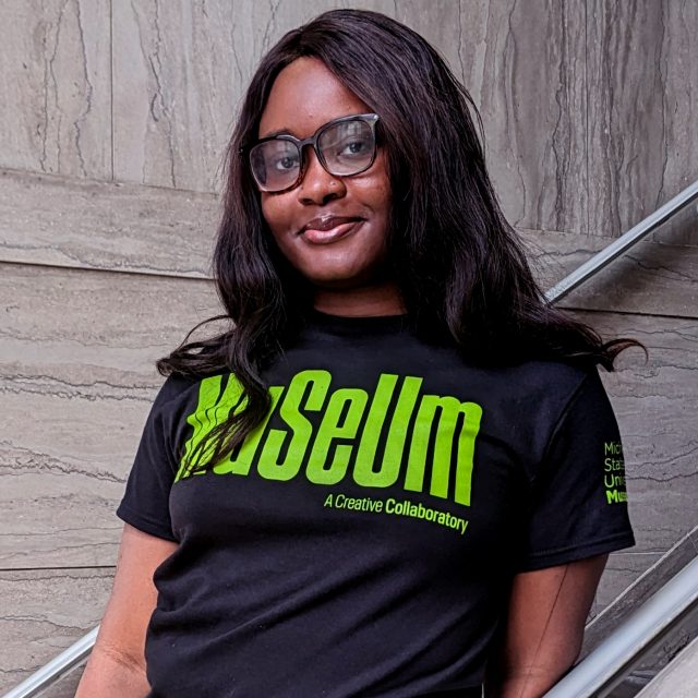 a young black woman with curly brown hair wearing a black t-shirt with green text reading "MUSEUM" stands in front of a marble staircase.