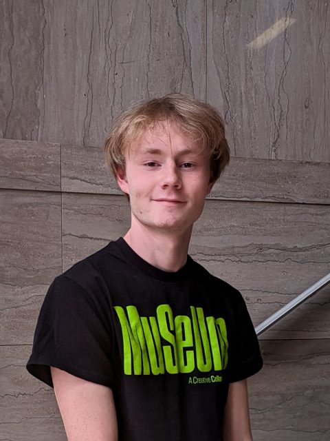 Block image A young MSU student with short blond hair standing on a marble staircase wearing a black shirt with green writing that reads 
