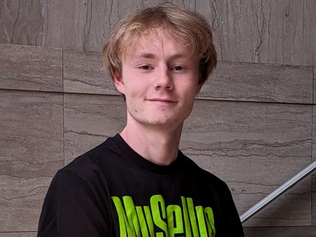 A young MSU student with short blond hair standing on a marble staircase wearing a black shirt with green writing that reads "Museum"