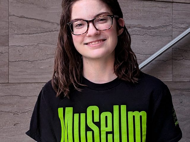 A young woman in glasses with wavy dark brown hair, wearing a black t-shirt with bright green text that reads "Museum A Creative Collaboratory"