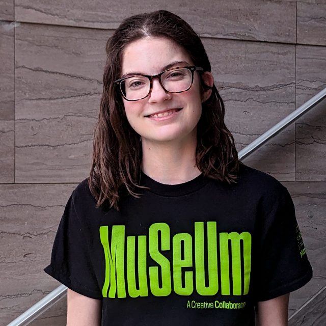 A young woman in glasses with wavy dark brown hair, wearing a black t-shirt with bright green text that reads "Museum A Creative Collaboratory"