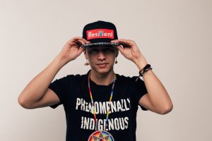 The image shows Frank Waln wearing a black cap that has the word "Resilient" in bold red and white letters on the front. Waln is holding the cap's brim with both hands, and dressed in a black t-shirt that says "PHENOMENALLY INDIGENOUS" in white text. Waln is also wearing a beaded necklace with a multicolored circular pendant, along with various beaded bracelets and earrings, including a small bear-shaped earring. The expression is neutral, and the background is a plain, light-colored wall. photo credit: Leslie Frempong