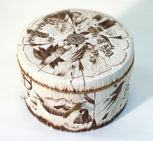 A round, decorative basket with intricate designs. The lid features detailed quill artwork of various natural elements, including birds, leaves, and flowers, arranged in a circular pattern. The basket is tied with a small piece of twine, giving it an organic, handcrafted appearance.