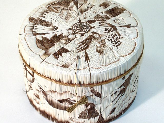 A round, decorative basket with intricate designs. The lid features detailed quill artwork of various natural elements, including birds, leaves, and flowers, arranged in a circular pattern. The basket is tied with a small piece of twine, giving it an organic, handcrafted appearance.