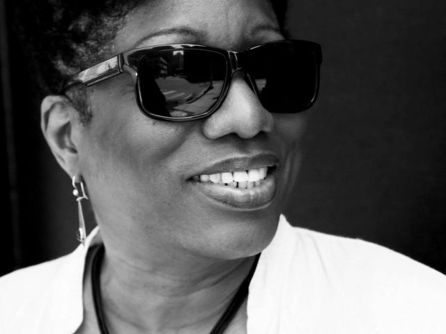 "A black-and-white portrait of Stacy "Hotwaxx" Hale wearing dark sunglasses and smiling confidently. They have short, styled hair and are dressed in a light shirt with a simple necklace. The background is solid and dark, drawing attention to their expressive face.