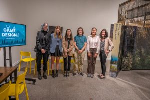 A group of six people stands together in an exhibition space, smiling at the camera. They are in front of a display featuring a forest scene and a sign reading "Boreal Forest / Écosystème boréal." To the left, a screen displays "GAME DESIGN: A Boreal Balance," along with additional text. Bright yellow chairs and tables are visible in the background, creating a modern and inviting setting. The group is diverse in terms of age, gender, and attire, ranging from casual to business casual.