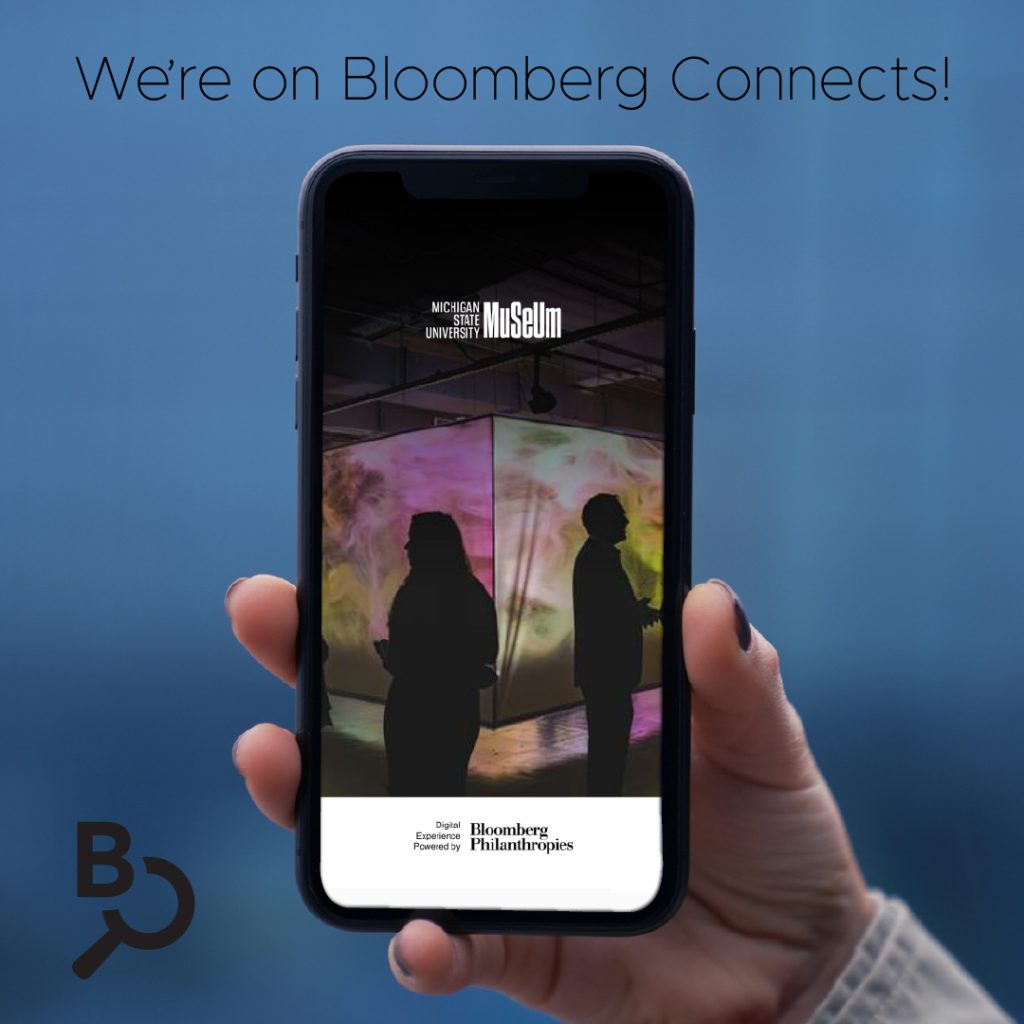 A tan hand with black nail polish holds up a smartphone against a dark blurry background. Visible on the phone is the opening image of the MSU Museum's Bloomberg Connects app, showing the MSU Museum logo and an image of silhouetted individuals standing in front of a brightly colored screen depicting space.