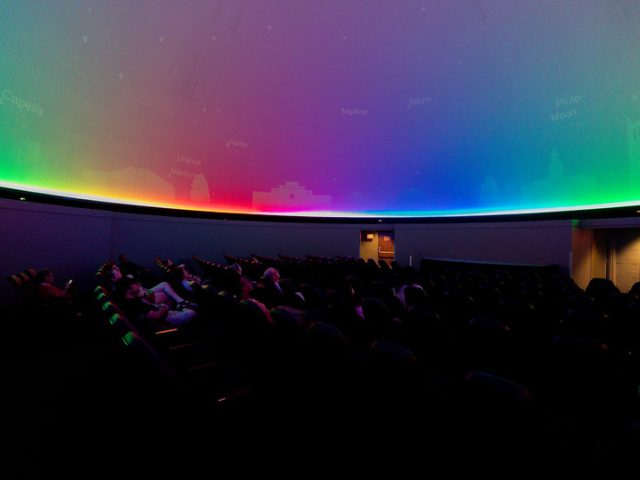 A planetarium with a large, domed ceiling illuminated by a gradient of rainbow colors, transitioning from red to green to blue. Faintly visible labels of celestial objects, such as 'Jupiter,' 'Neptune,' and 'Saturn,' are projected onto the dome. Audience members are reclining in dark theater seats, gazing at the display.