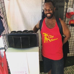 A man wearing a red tank top with 'Club Heaven' and a palm tree graphic in yellow, stands next to a black speaker on a white pedestal. He carries a backpack and is in a booth with various merchandise, including hats and shirts on a grid wall display.